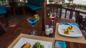A wooden table with a white plate of food and a blue cocktail, set on a terrace with a view of the ocean. - Blue Ocean Resort (Mui Ne)