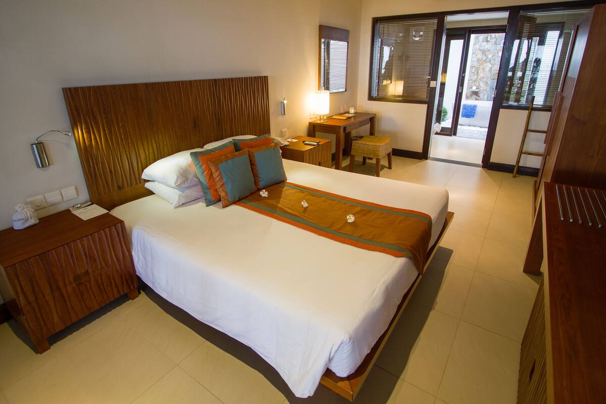 A hotel room with a large bed, a wooden headboard, a bedside table with a lamp, a small table, and a view of the outside through a sliding door.
