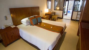 Habitación de hotel con una cama grande, cabecera de madera, mesita de noche con lámpara, una pequeña mesa y vistas al exterior mediante una puerta corredera.