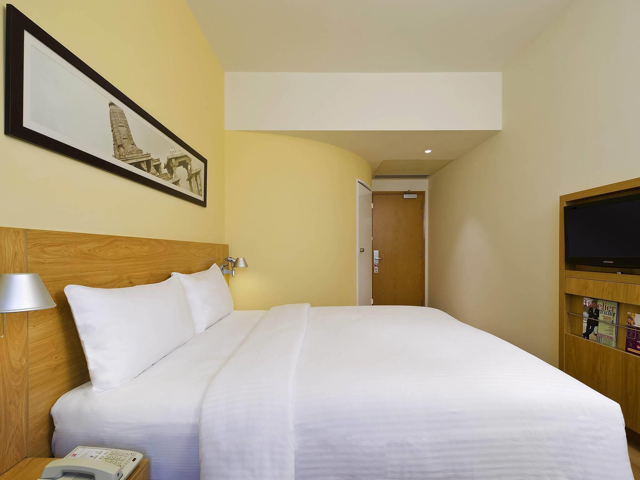 A hotel room with a bed, a bedside lamp, a telephone, a television, and a framed picture on the wall.