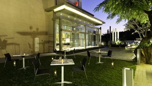 An outdoor dining area with tables and chairs, a glass building with interior lighting, and a well-maintained lawn. - ibis Nashik Hotel (Nashik)