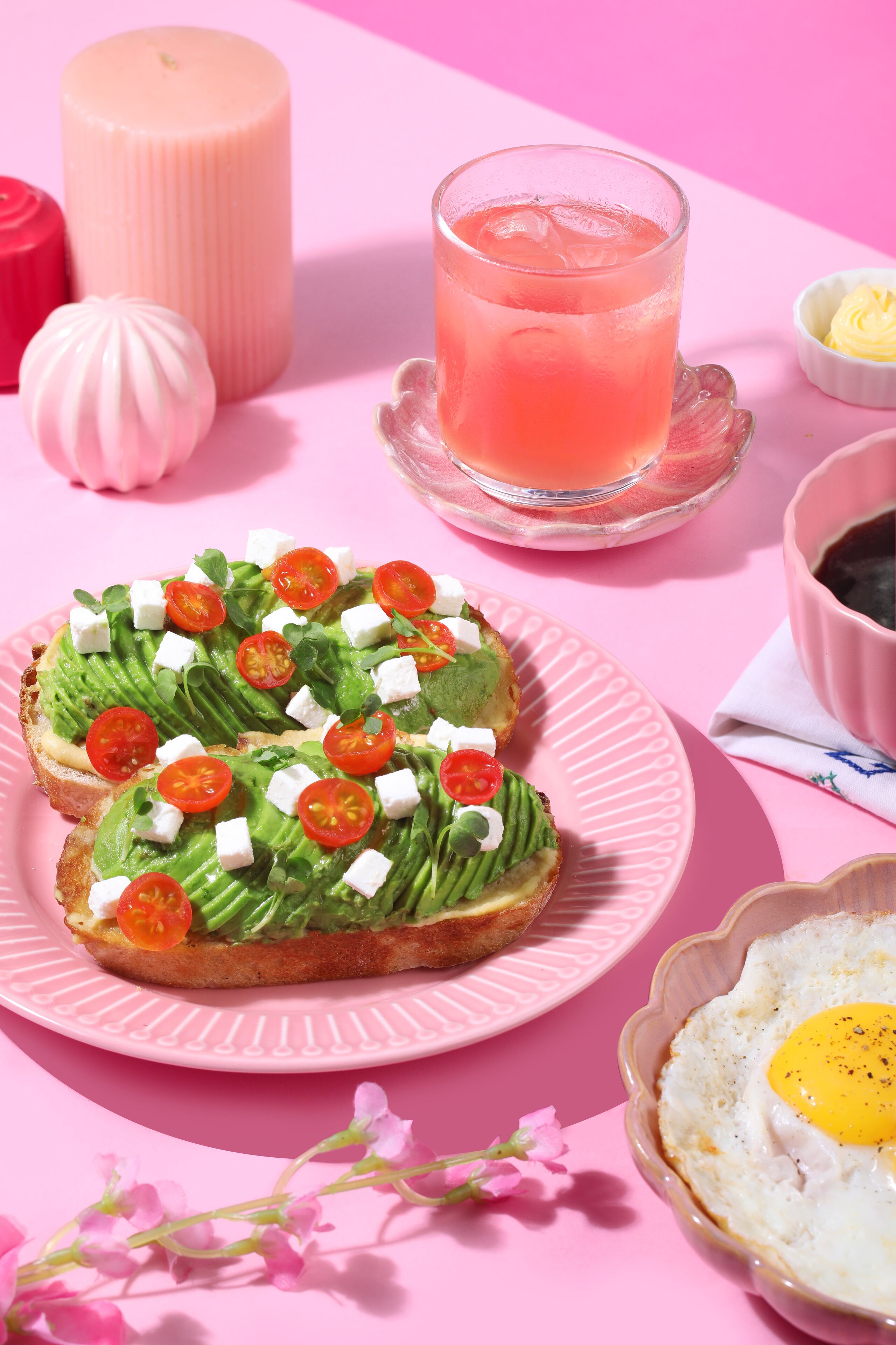 A pink table with a plate of avocado toast, a glass of pink drink, and a bowl of butter.