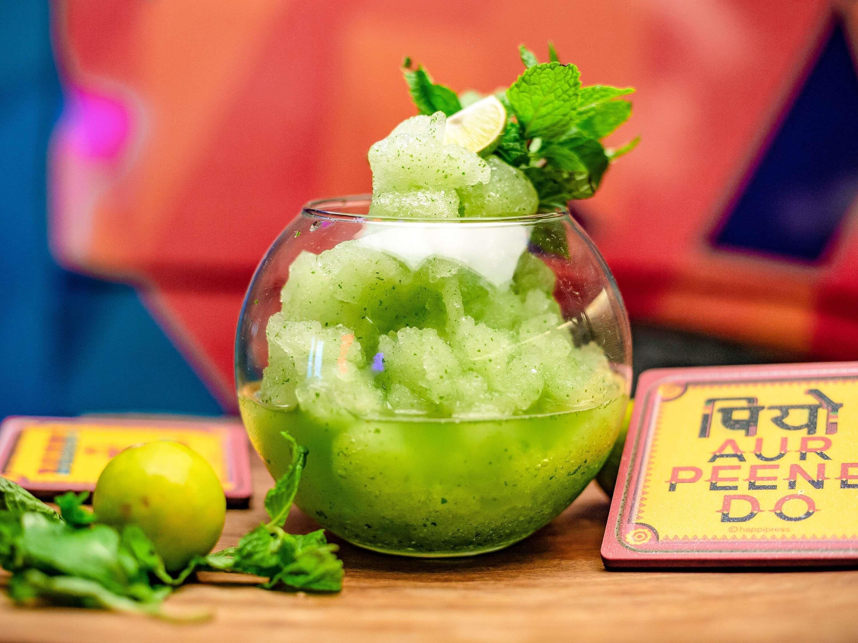 A glass bowl with a green beverage garnished with mint leaves and a lime slice, placed on a wooden surface with a colorful background.