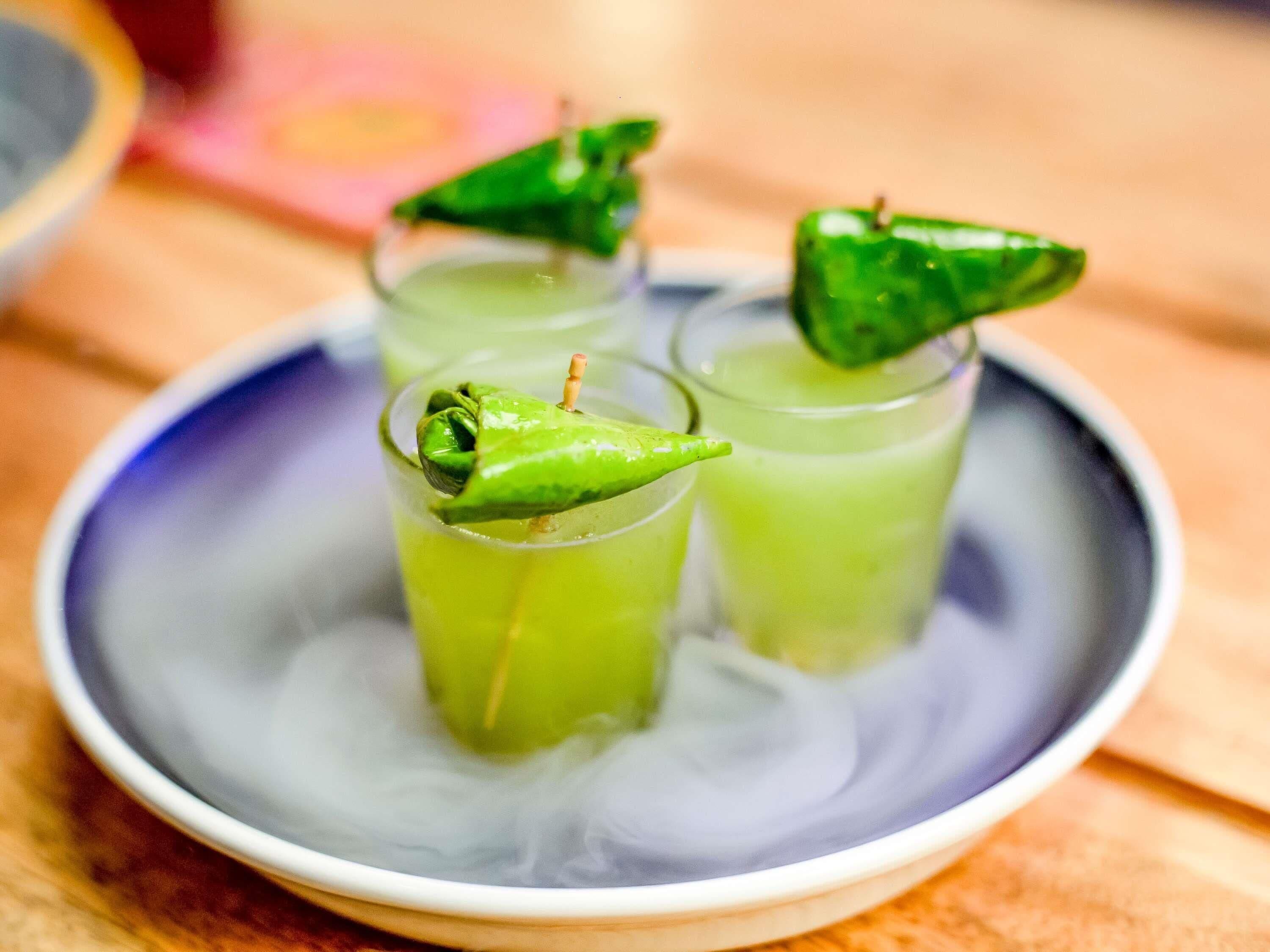 A bowl with three glasses of green liquid, each garnished with a green chili pepper.