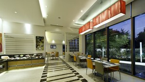 A modern restaurant with a long counter, dining tables, and chairs. There is a large window with a view of trees and a road. - ibis Nashik Hotel (Nashik)