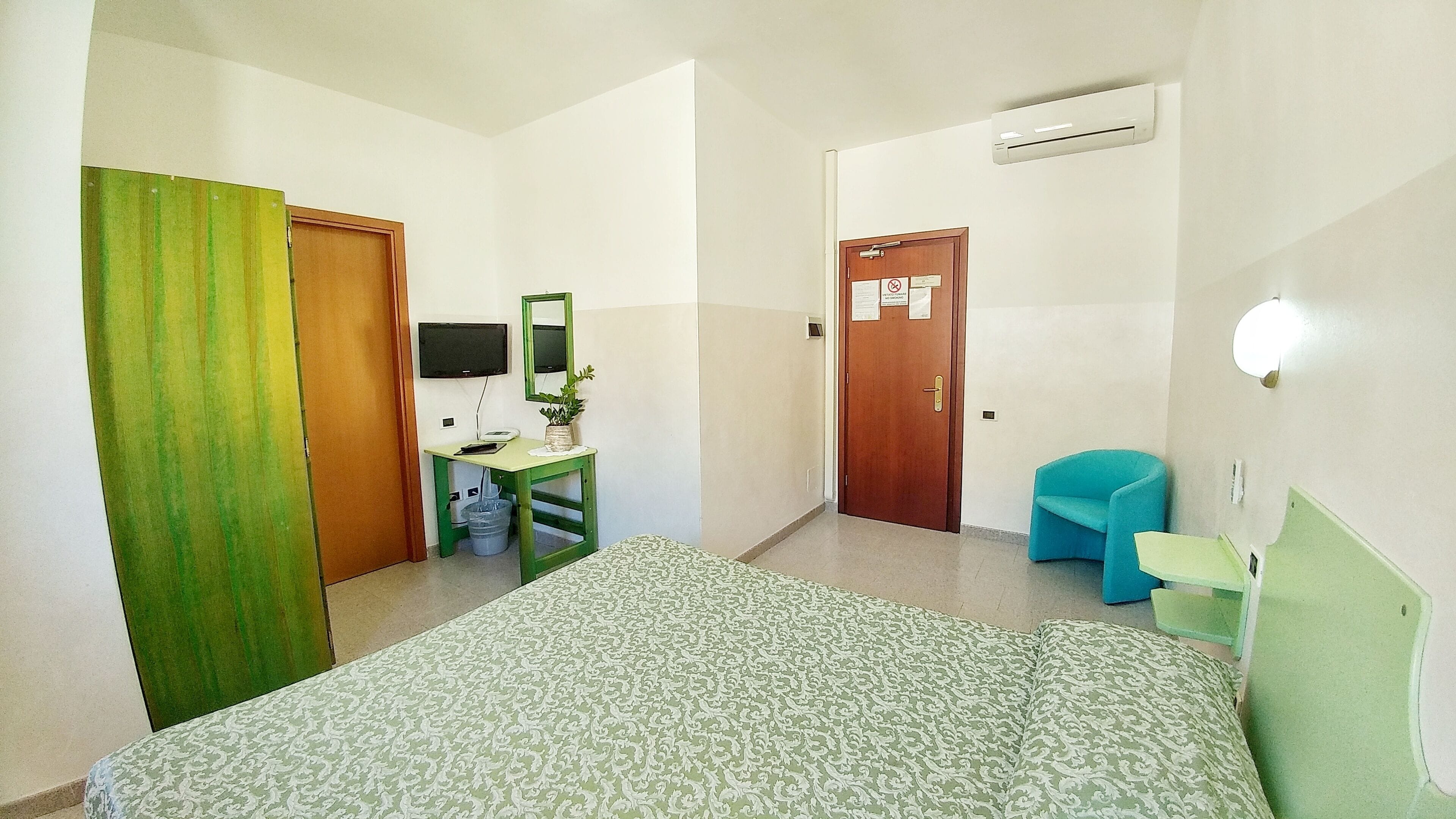 Una habitación de hotel con una cama, un escritorio con una computadora, una silla, una cortina verde y una puerta con un cartel.