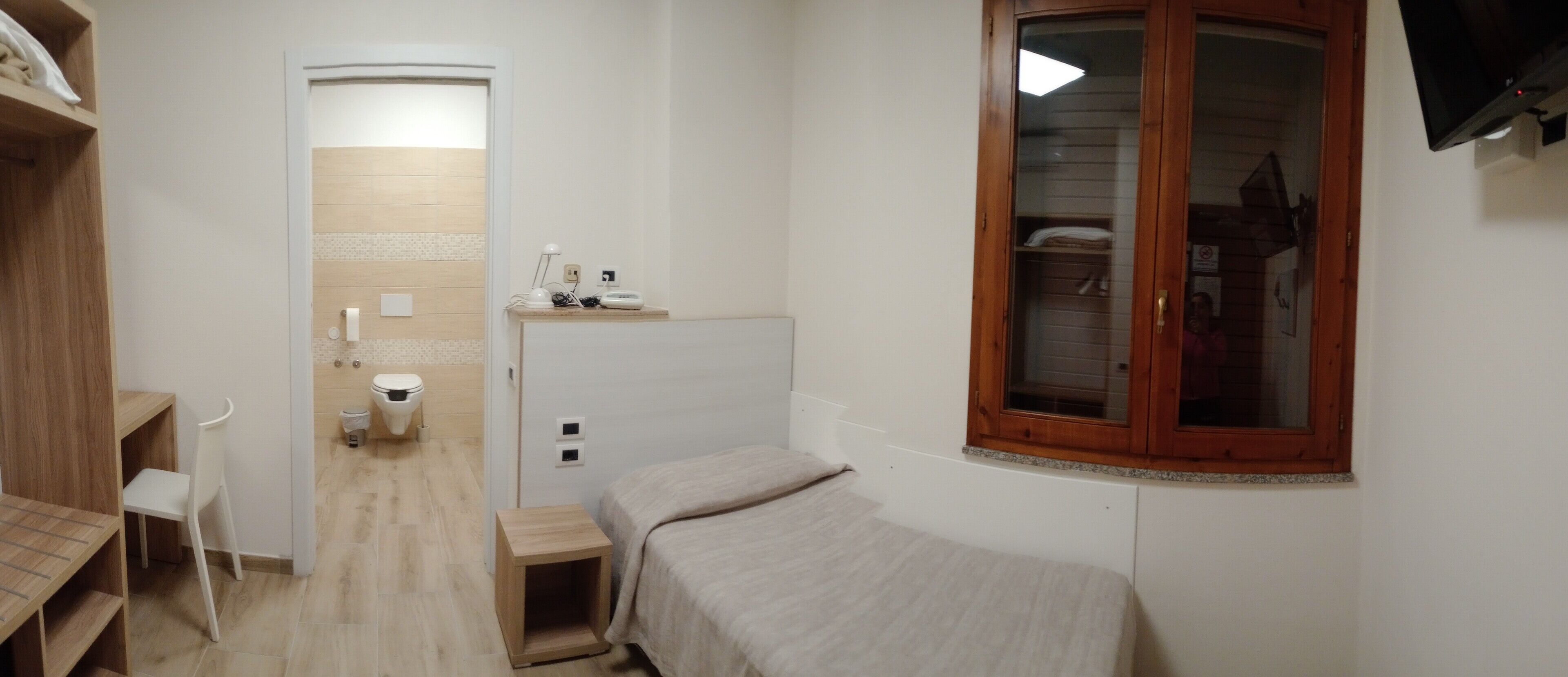 Une petite pièce, de taille modeste, comprenant un lit, un bureau et une chaise. Il y a une fenêtre avec un cadre en bois et un téléviseur fixé au mur.