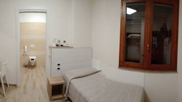 A small, compact room with a bed, a desk, and a chair. There is a window with a wooden frame and a television mounted on the wall.