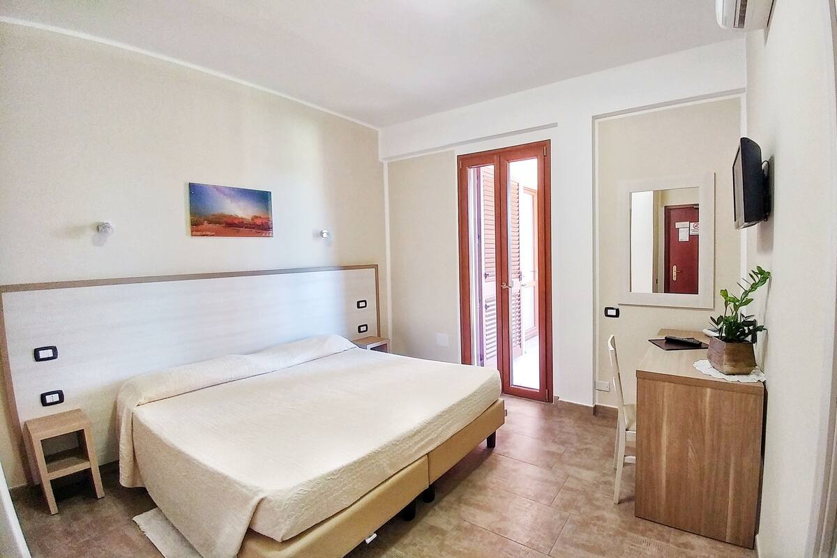 A hotel room with a bed, a small desk with a plant, a mirror, and a painting on the wall.