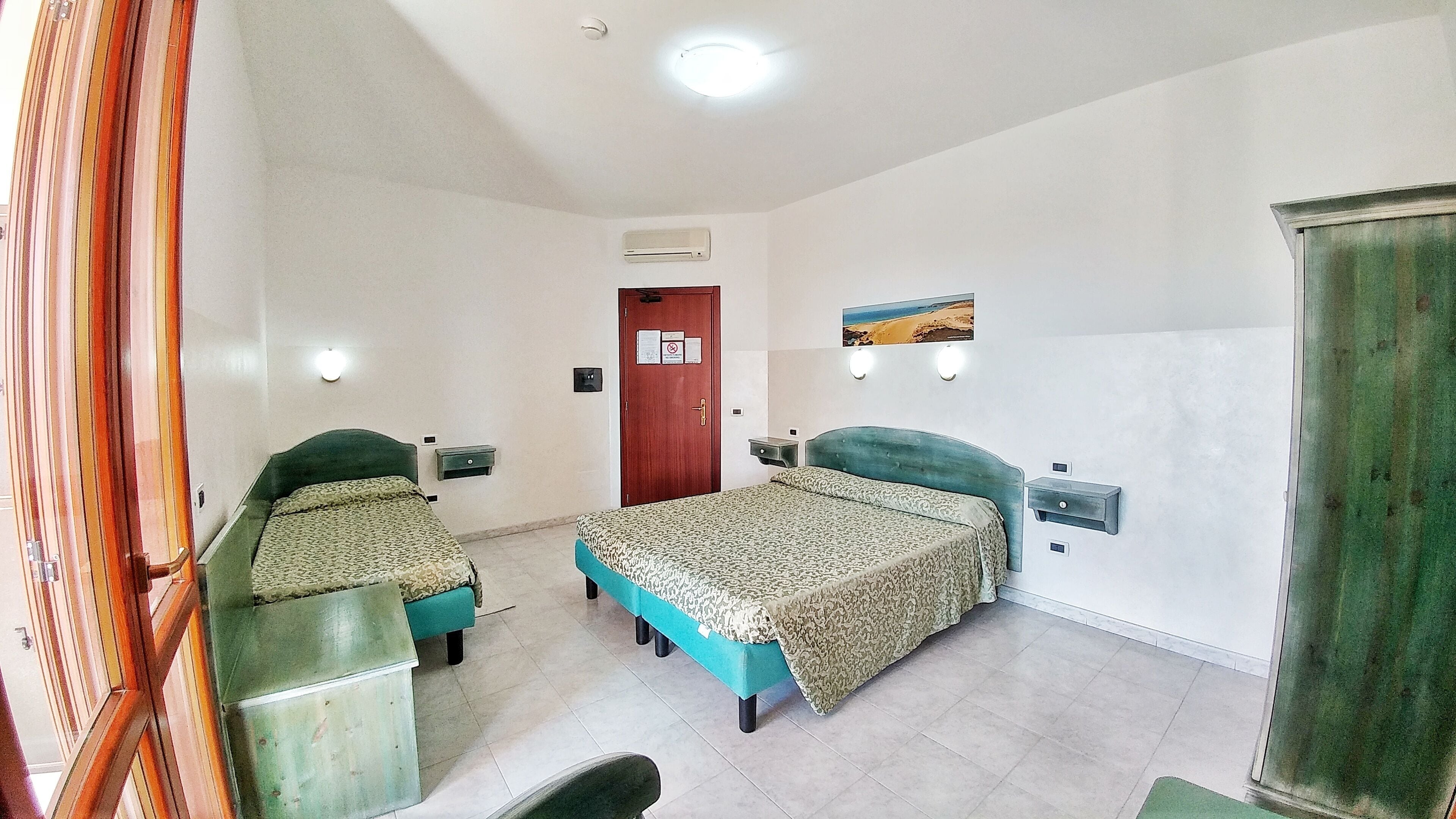 A hotel room with two single beds, a wooden wardrobe, a small table, a wall-mounted air conditioner, and a door with a notice.