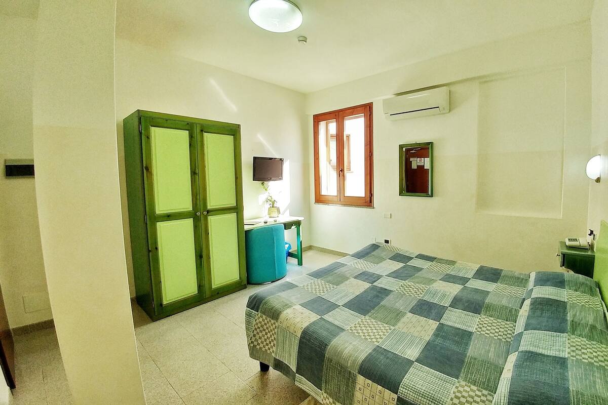 A bedroom with a bed, a desk, a wardrobe, a window, and an air conditioning unit.