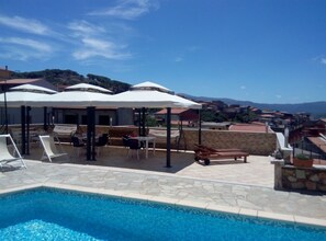 A poolside area with lounge chairs and umbrellas, a stone pool, and a view of a town with buildings and hills in the background.