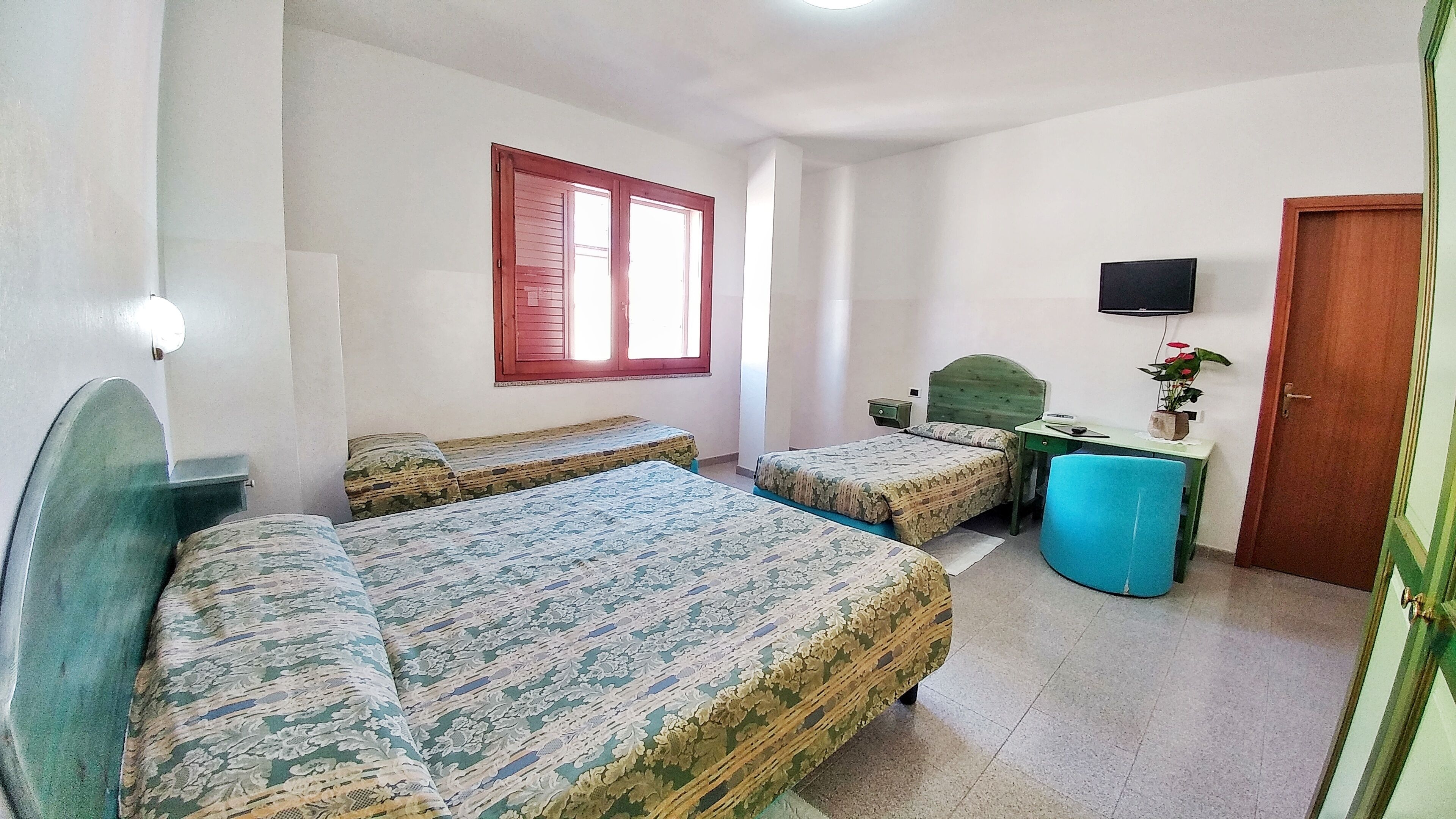 Quarto com duas camas, uma escrivaninha, uma televisão e uma janela com persianas vermelhas.