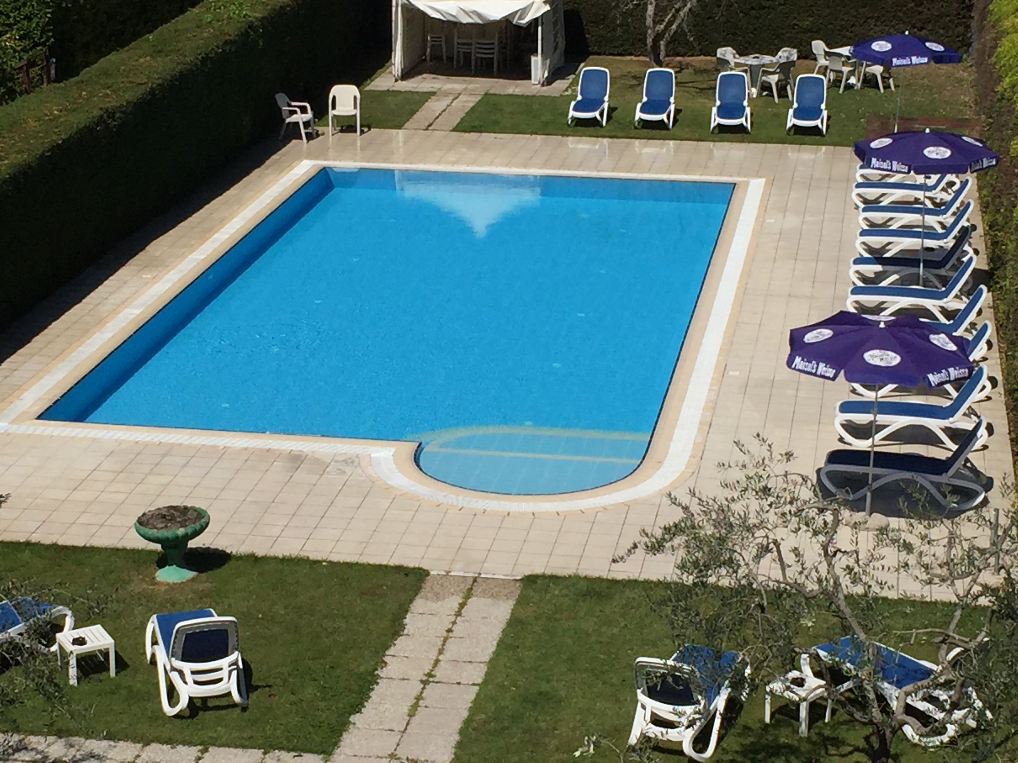 Et rektangulært swimmingpool med flisebelagte omgivelser, omgivet af liggestole og parasoller.