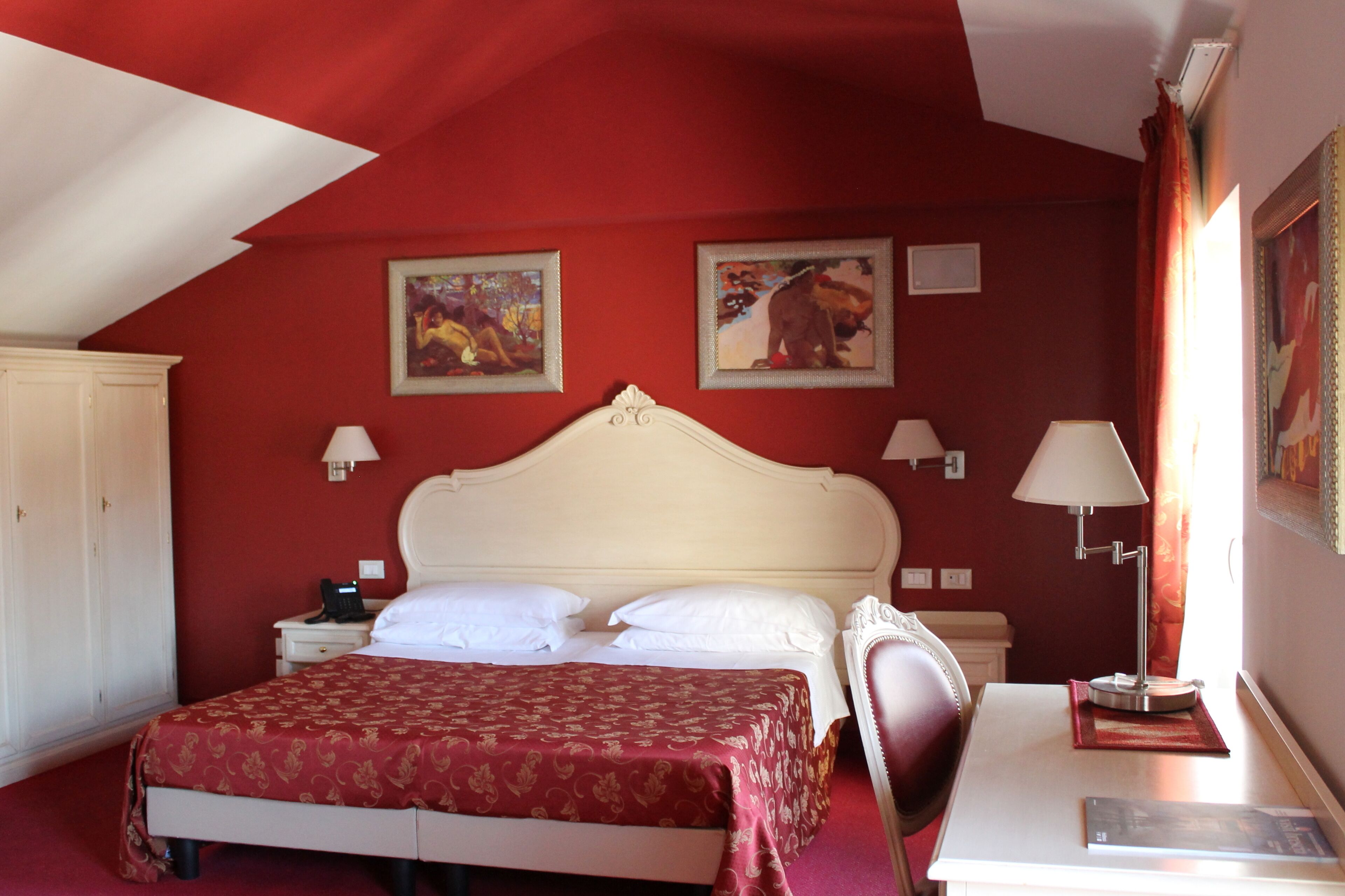 Ein Schlafzimmer mit roter Wand, einem weißen Bett, zwei Bildern, einem Schreibtisch mit Lampe und einem Sessel.