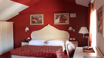 Ein Schlafzimmer mit roter Wand, einem weißen Bett, zwei Bildern, einem Schreibtisch mit Lampe und einem Sessel.