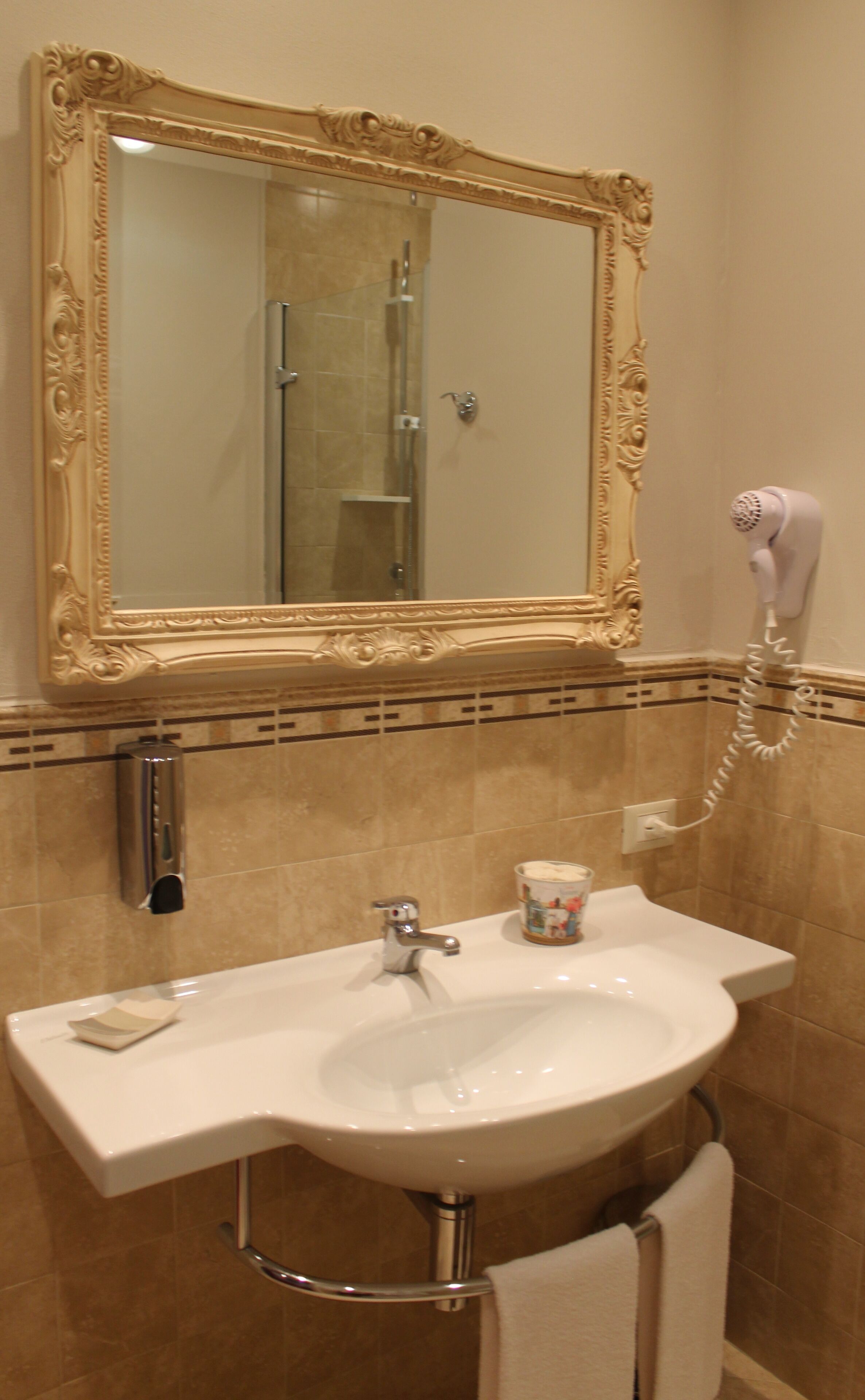 Un baño con un espejo grande y decorativo, un lavabo blanco, un secador de pelo de mano y una toalla colgada en un perchero.