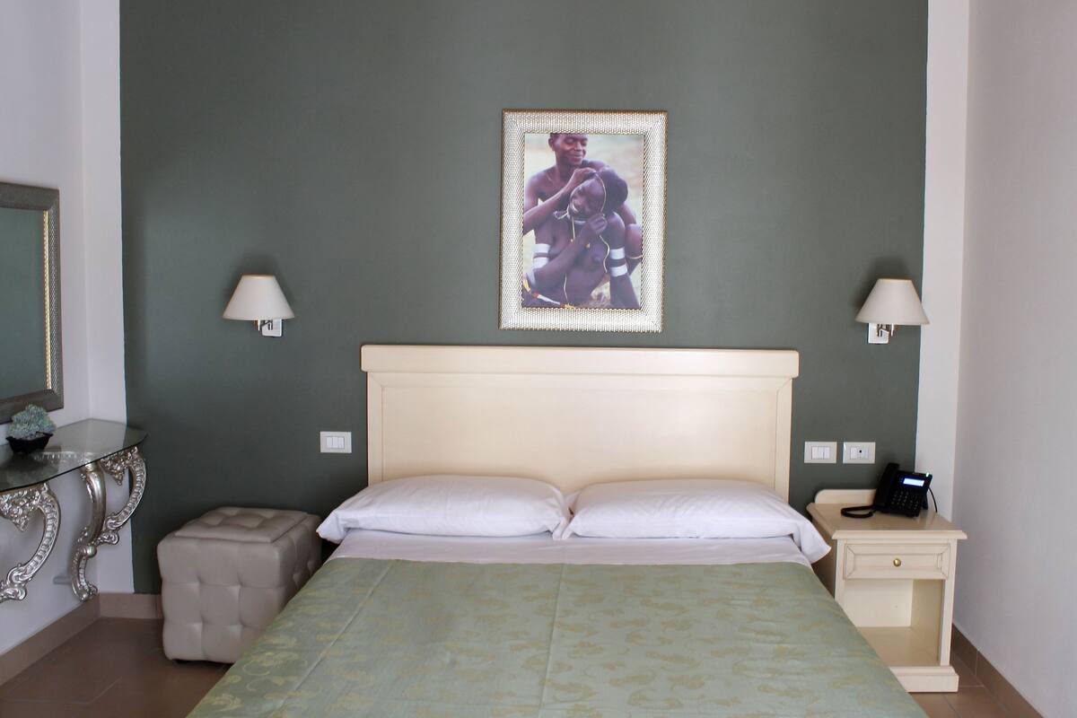 A bedroom with a bed, bedside lamps, a nightstand with a phone, a small ottoman, and a framed picture on the wall.