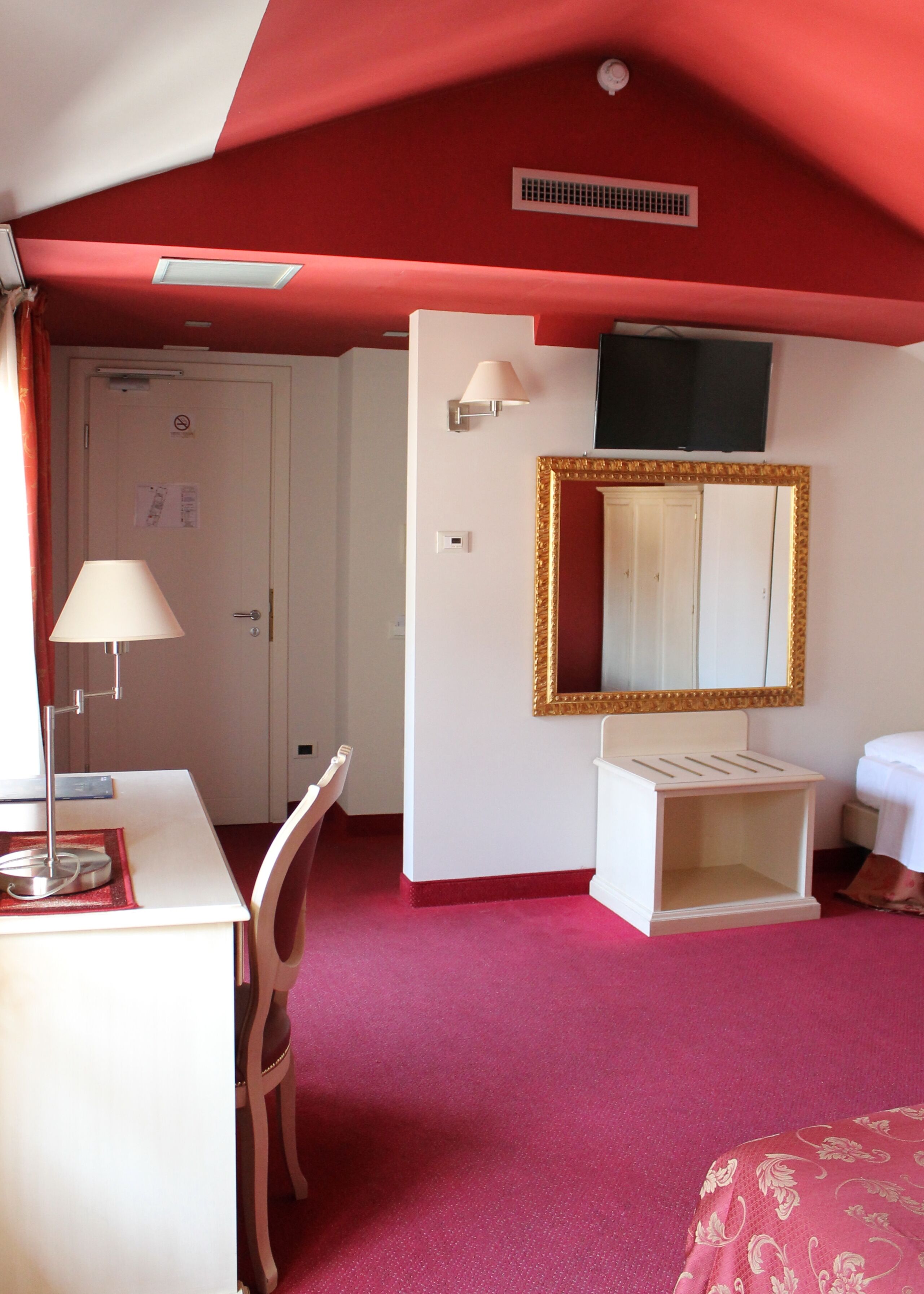 A room with a desk, chair, bedside table, and a large mirror with a gold frame.