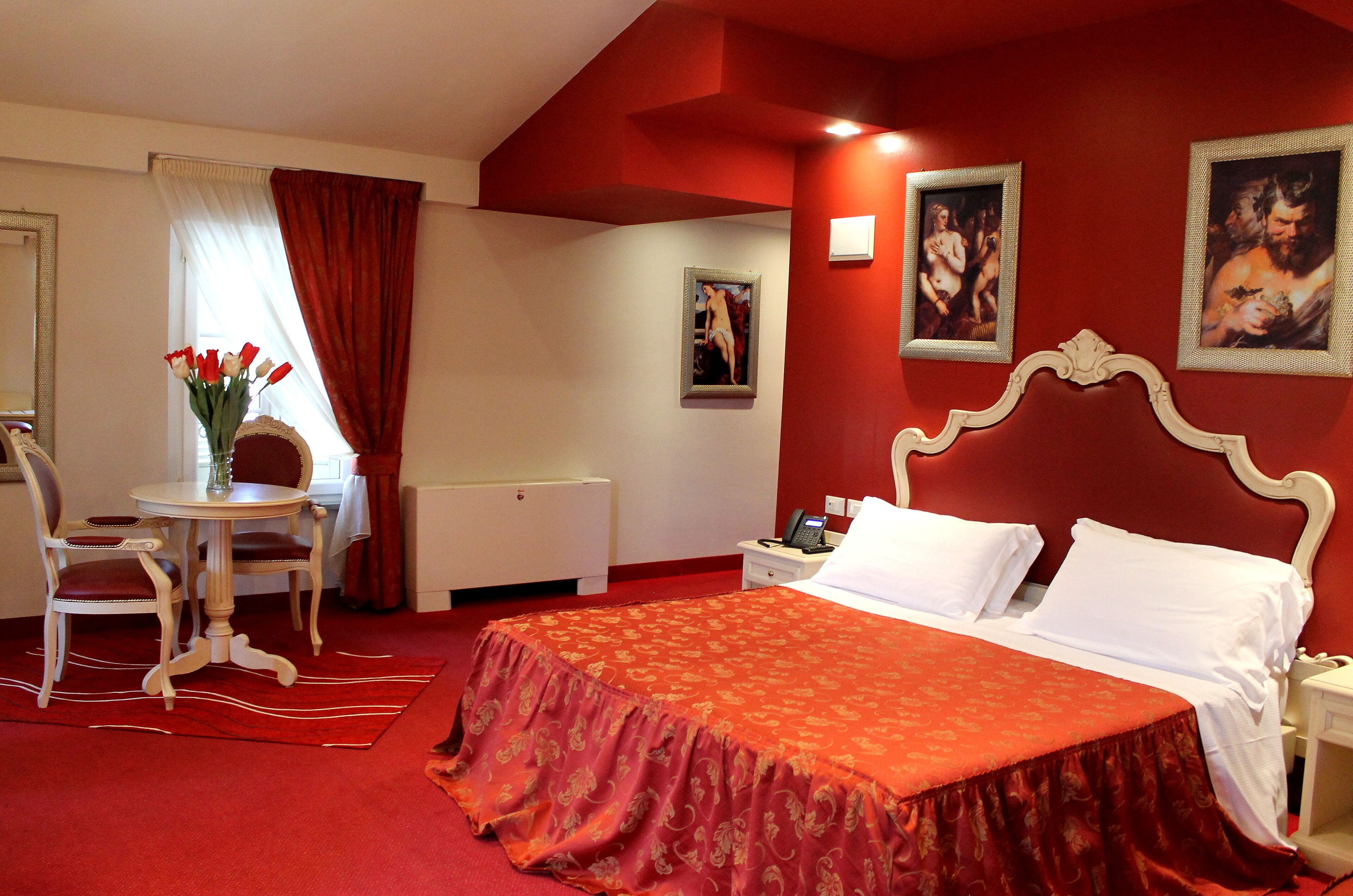 Habitación de hotel con alfombra roja, cama con ropa blanca, una mesita con un jarrón de flores rojas, una silla y dos cuadros en la pared.