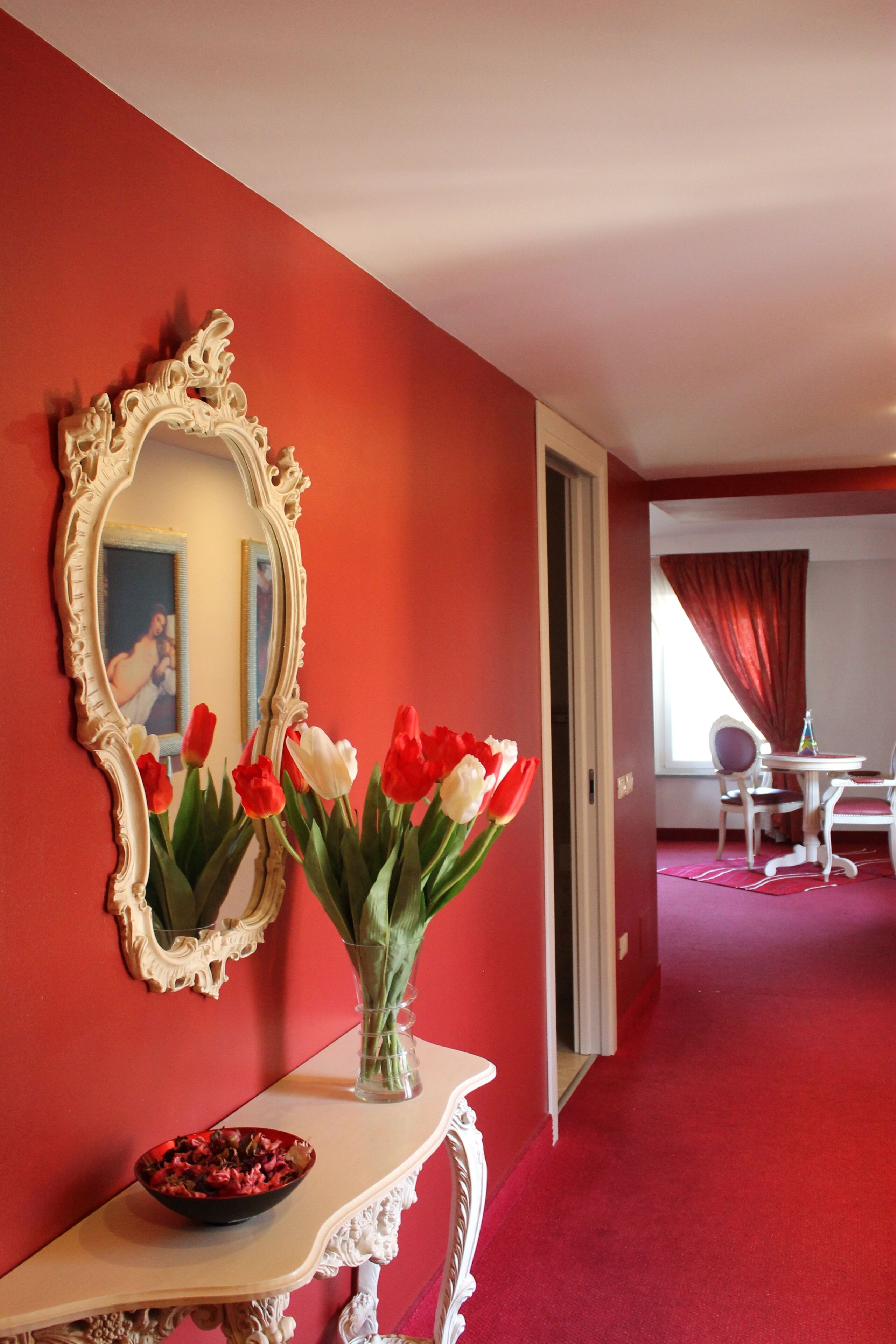 A room with a red wall, a white ornate mirror, a vase with red and white tulips, a small table with a bowl, and a dining area in the background.