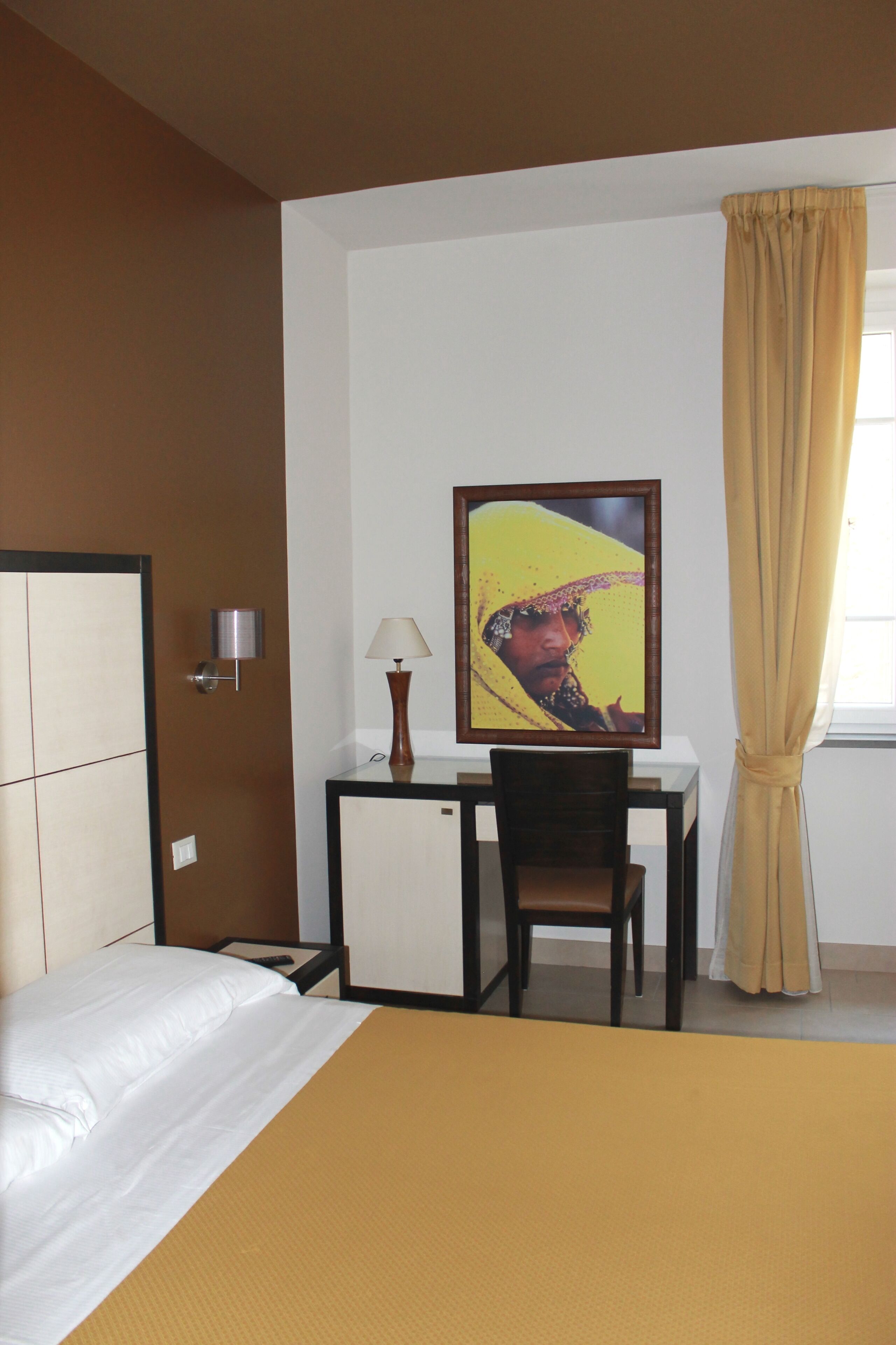A hotel room with a bed, a desk, a chair, a lamp, and a painting on the wall.