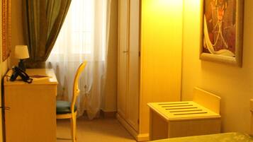 A hotel room with a bed, a desk with a lamp, a chair, a wardrobe, and a painting on the wall.
