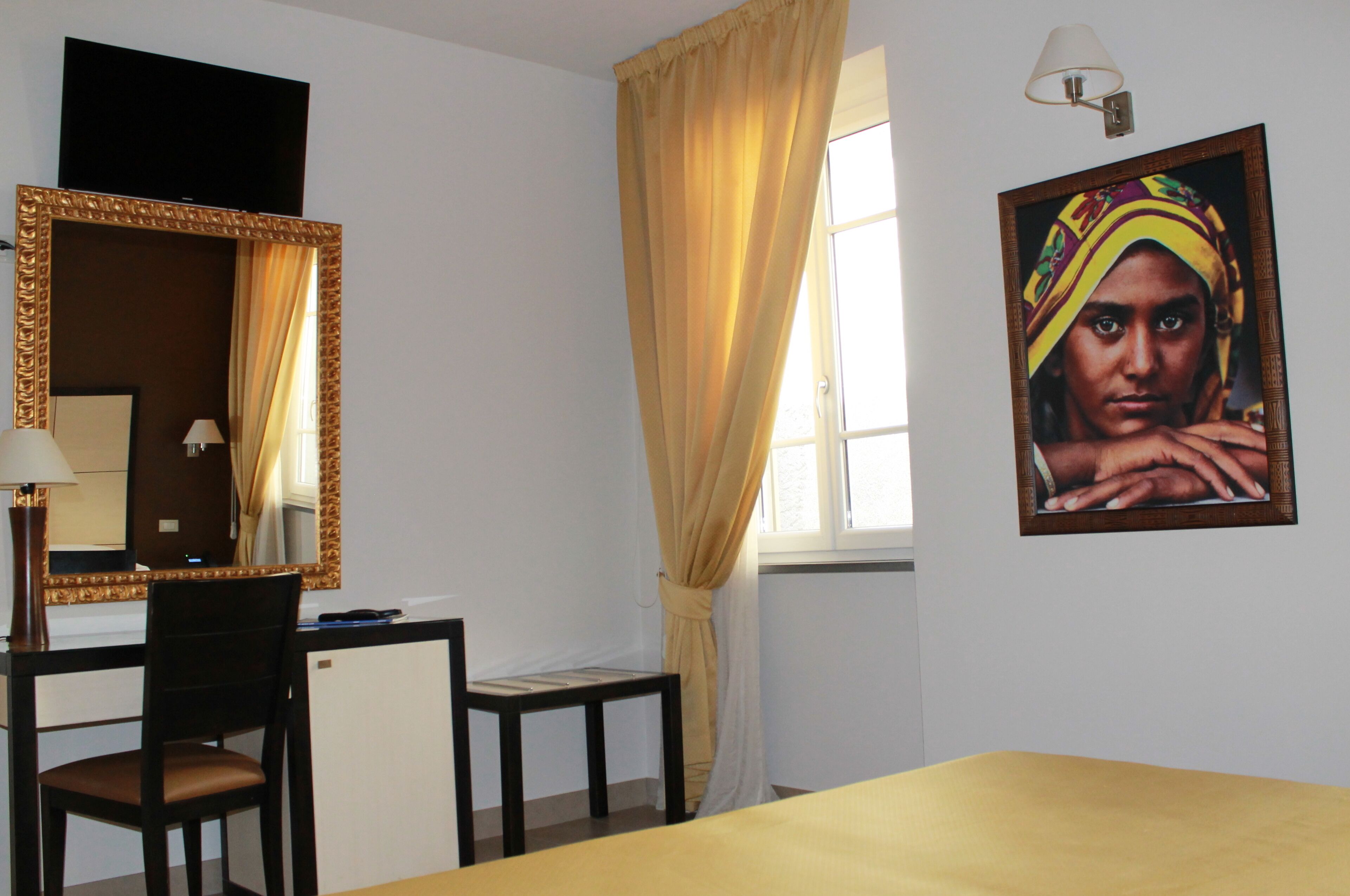 Habitación de hotel con escritorio, silla, espejo, ventana con cortinas y un cuadro en la pared.