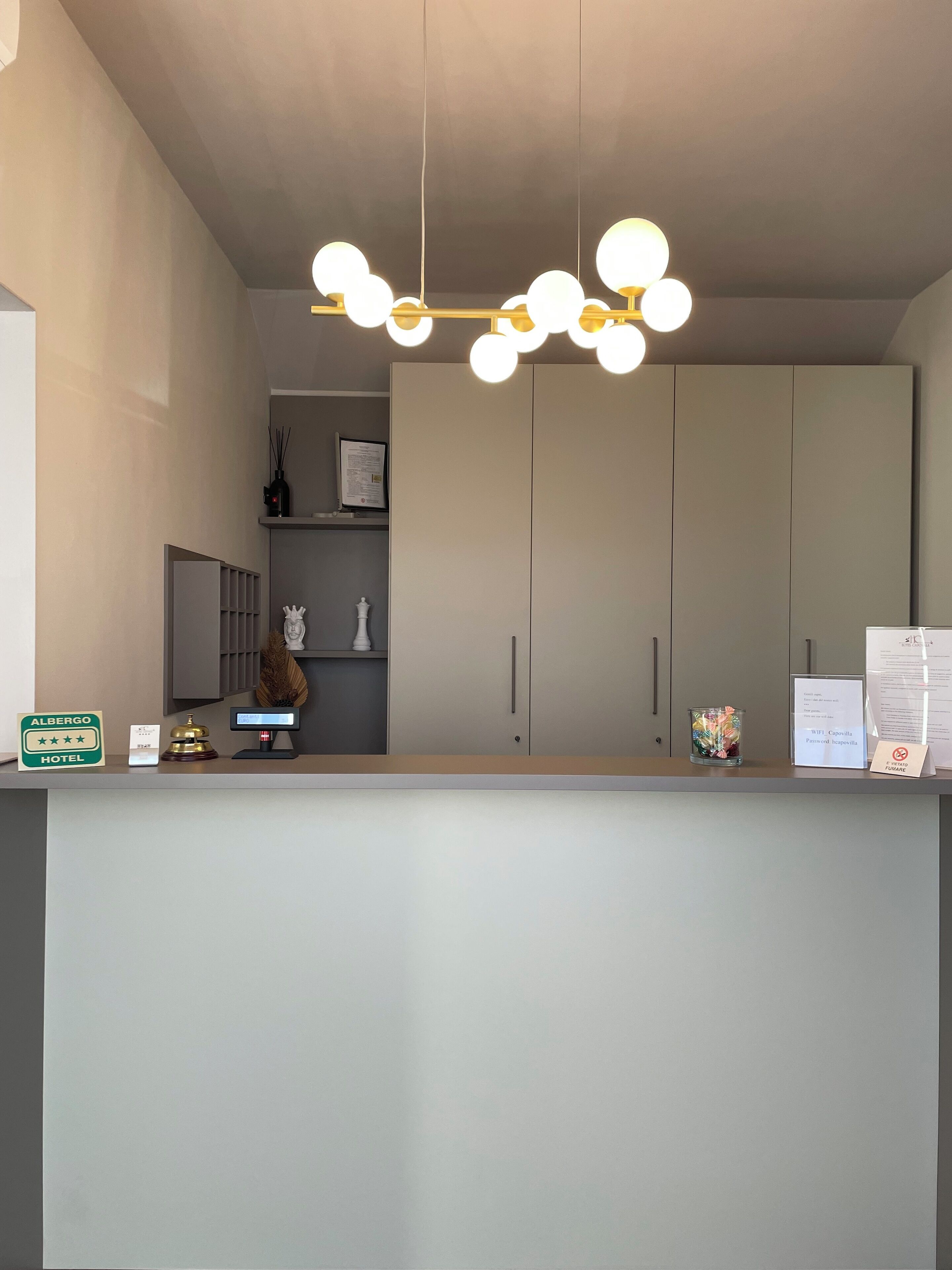 Un comptoir d’accueil d’hôtel au design moderne, comprenant un luminaire avec plusieurs ampoules sphériques, un comptoir de réception, un certificat encadré et un petit objet décoratif.