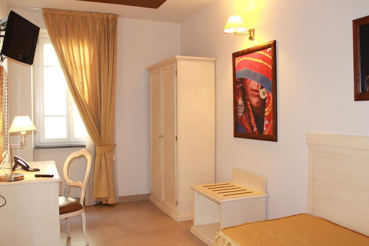 A bedroom with a bed, a desk with a lamp, a chair, a wardrobe, and a painting on the wall.