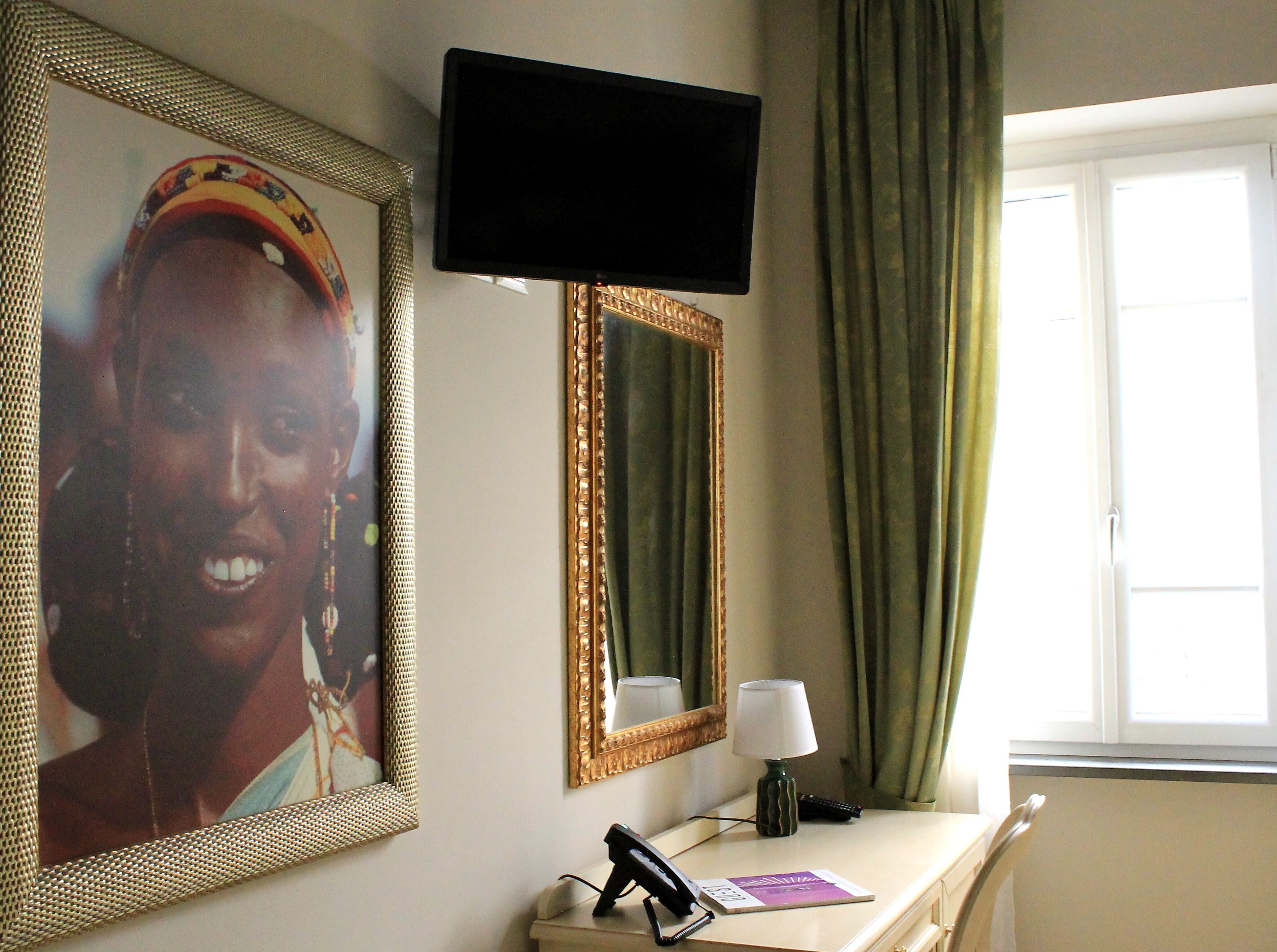 A hotel room with a television, a framed picture, a mirror, a lamp, a desk with a phone and a book, and a window with curtains.