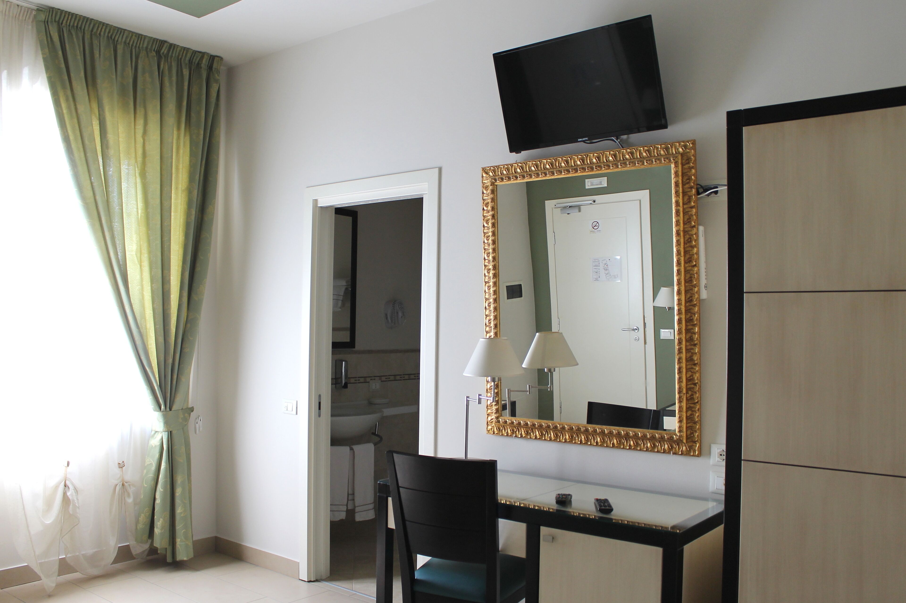 Une chambre d’hôtel équipée d’un bureau, d’une chaise, d’une télévision, d’un miroir et d’une porte donnant accès à une salle de bain.