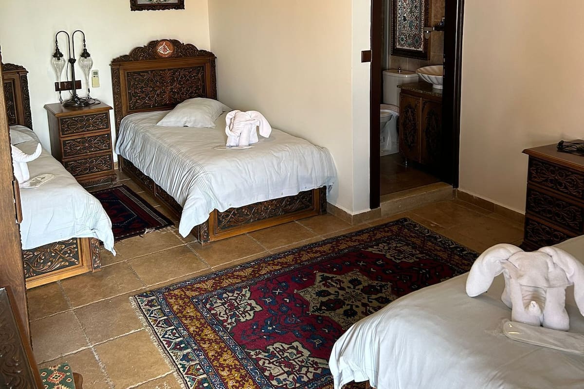 Habitación tradicional con dos camas, techo de madera, alfombra estampada y puerta que da a un baño.