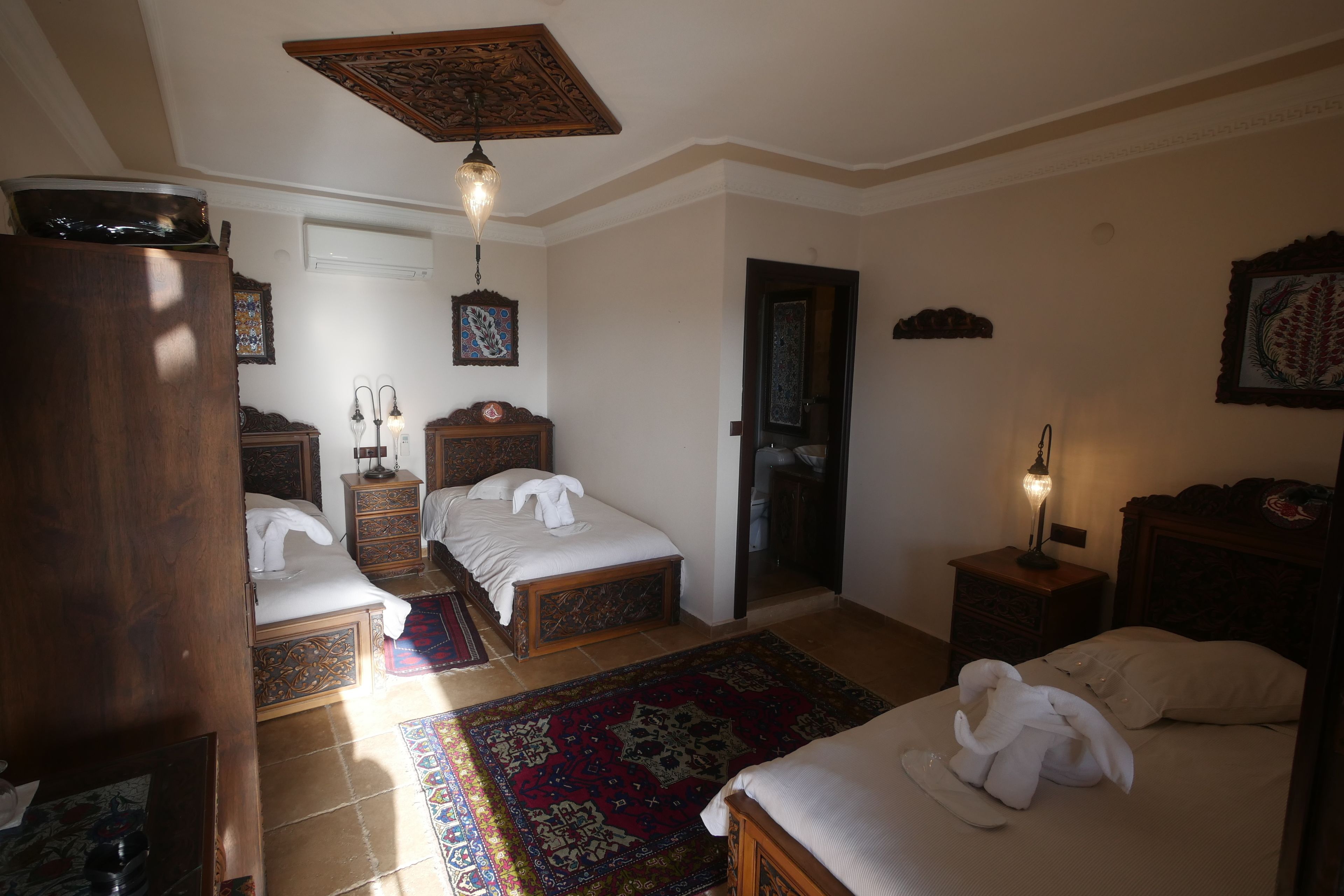 A hotel room with two beds, a wooden wardrobe, a nightstand with a lamp, and a patterned rug.