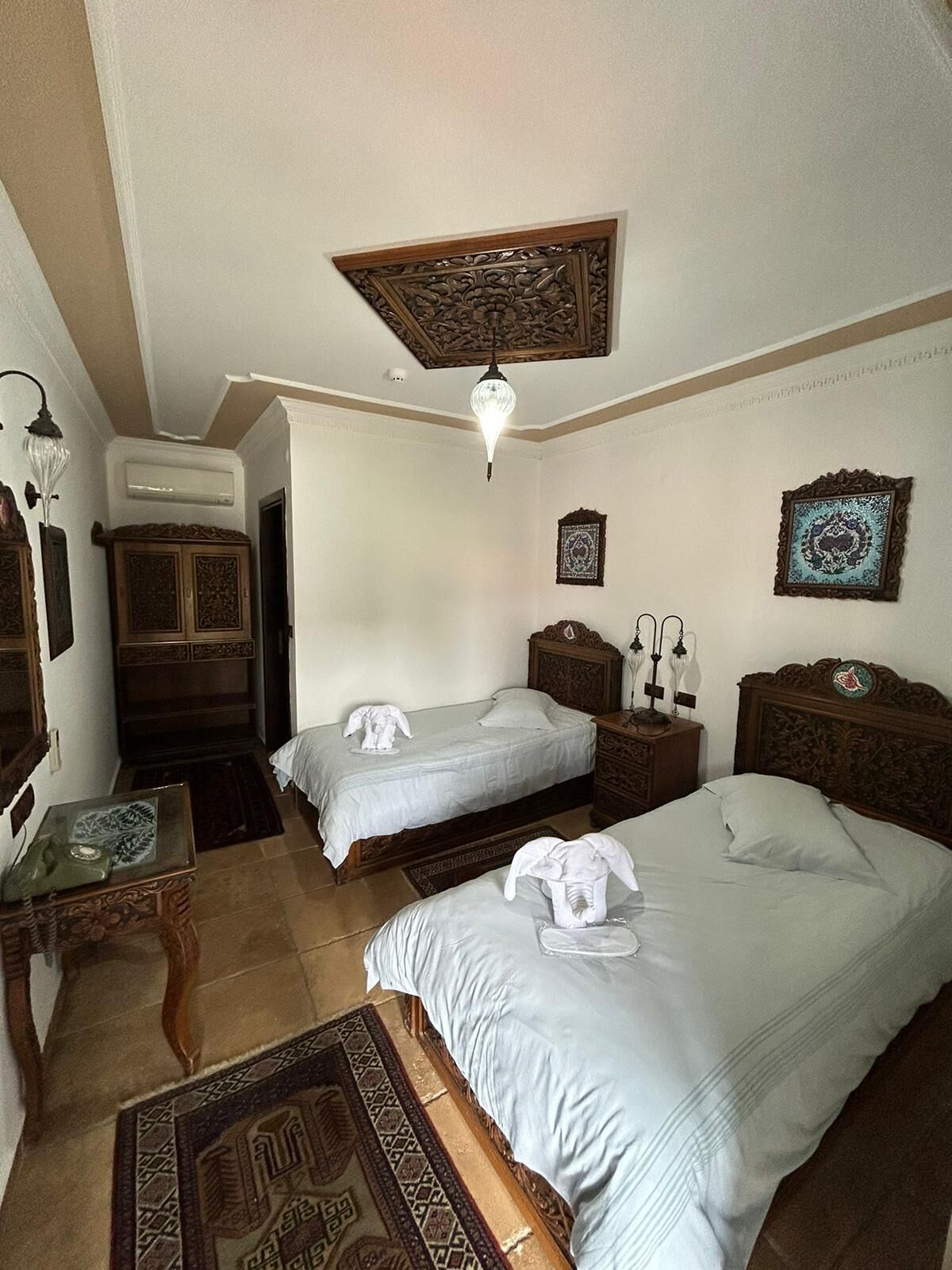 A hotel room with two beds, a wooden headboard, a nightstand with a lamp, a decorative wall hanging, and a patterned rug.