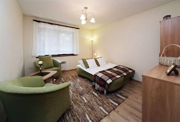 Un dormitorio con cama, un sillón verde, un escritorio de madera, una lámpara y una ventana con cortinas.