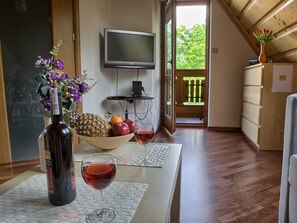 A room with a wooden floor, a TV mounted on the wall, a table with a bottle of wine, a bowl of fruit, and a vase of flowers.