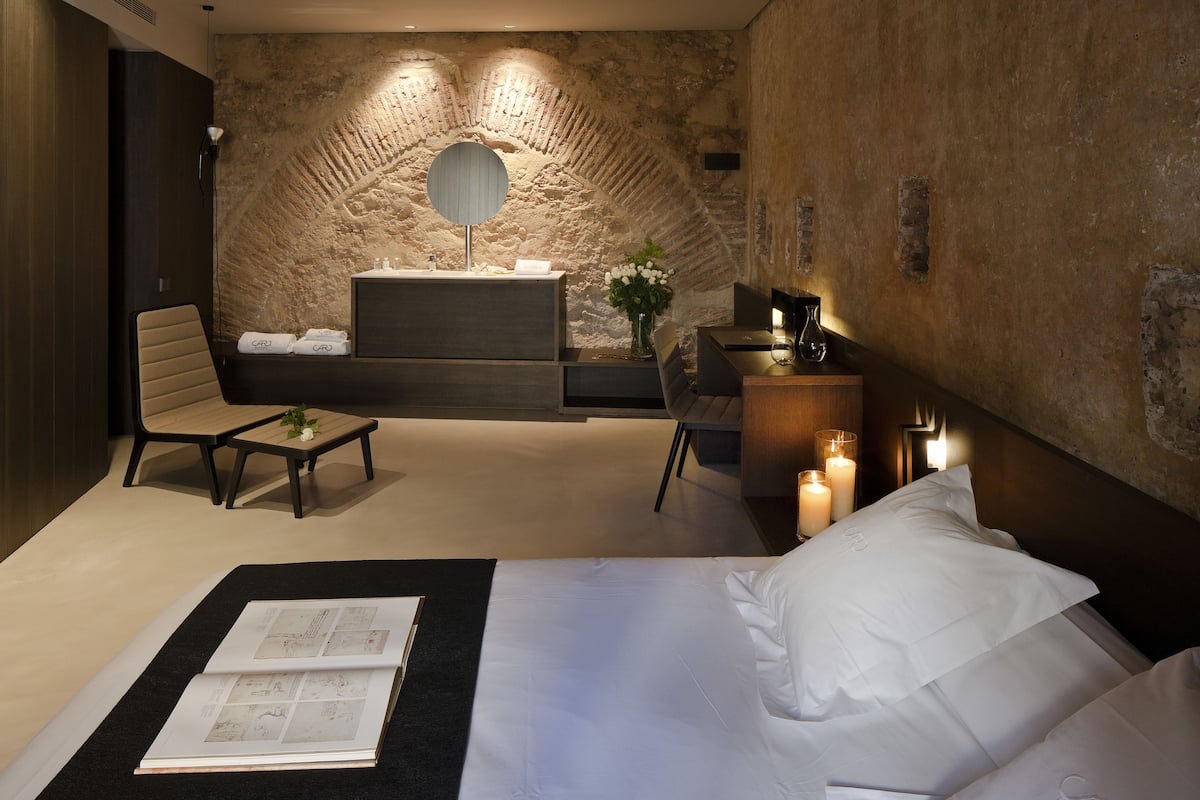 Un dormitorio moderno con una pared de acento de piedra, un espejo redondo, una cama con ropa blanca, un escritorio con un florero y una silla.