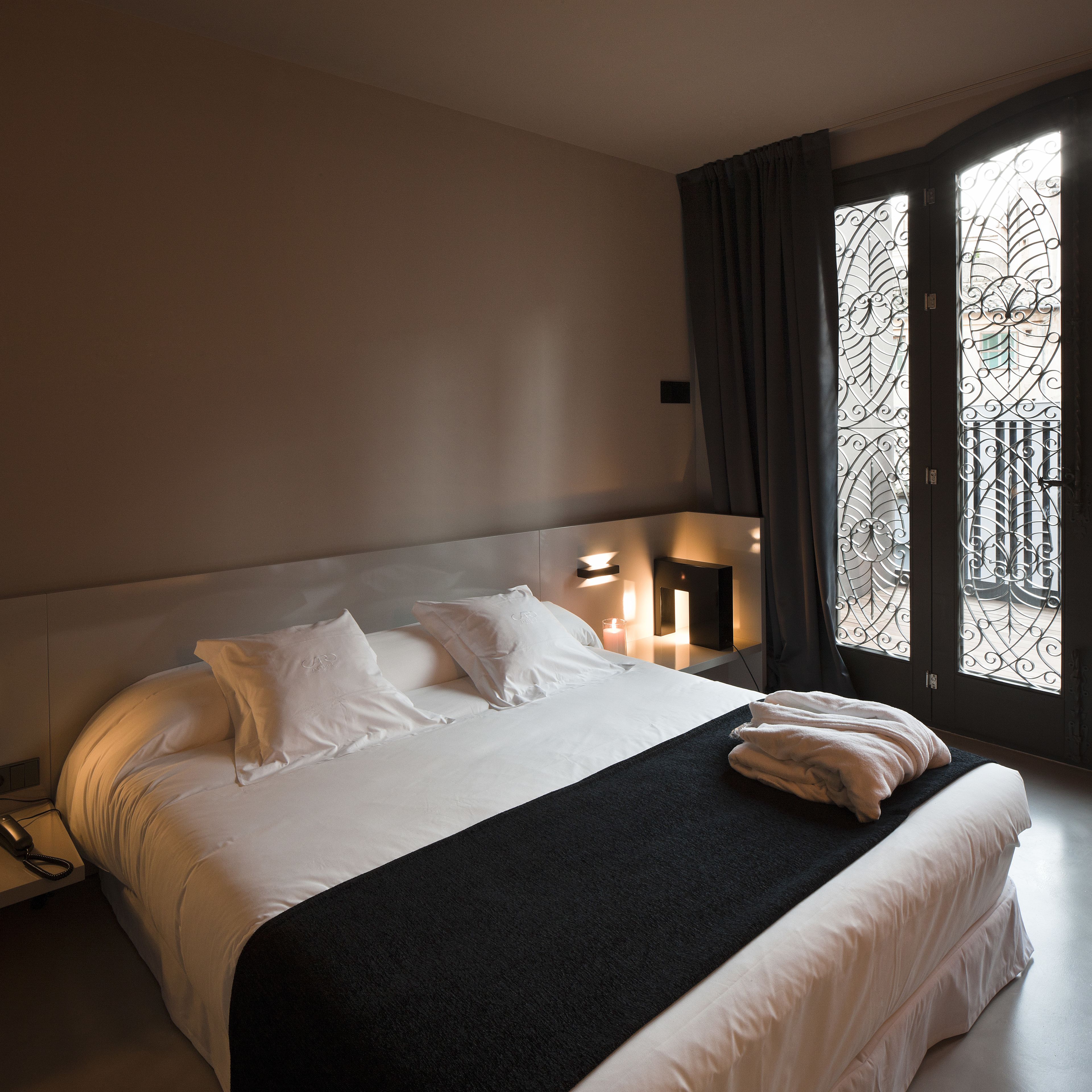 A hotel room with a large bed, a bedside table with a lamp, and a view through a window with decorative iron bars.