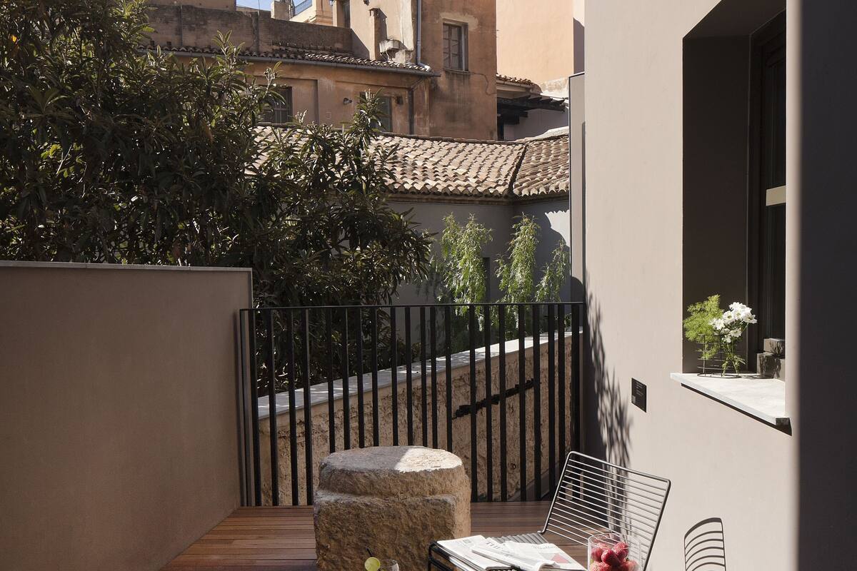Una terraza de madera con una mesa y dos sillas, una mesita redonda con un frutero, y una ventana con una planta.