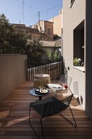 Una terraza de madera con una mesa y dos sillas, una mesita redonda con un frutero, y una ventana con una planta.