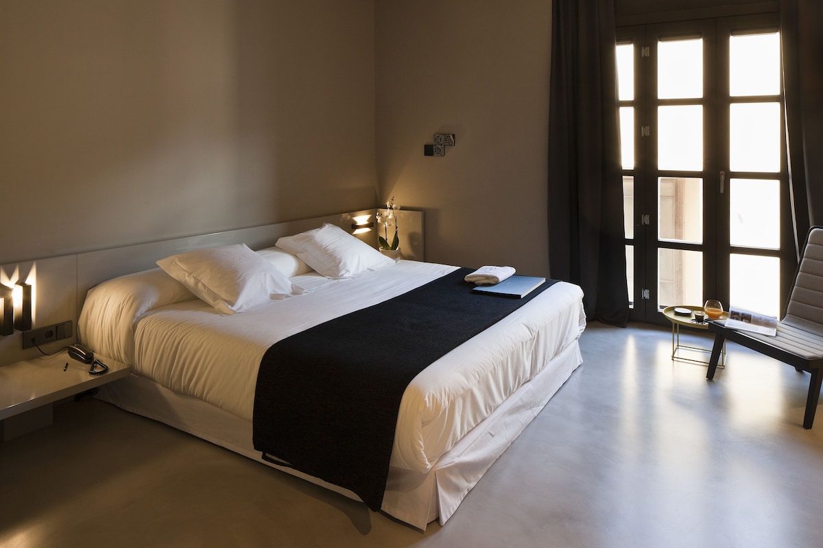 A hotel room with a large bed, bedside tables, a chair, and a window with curtains.