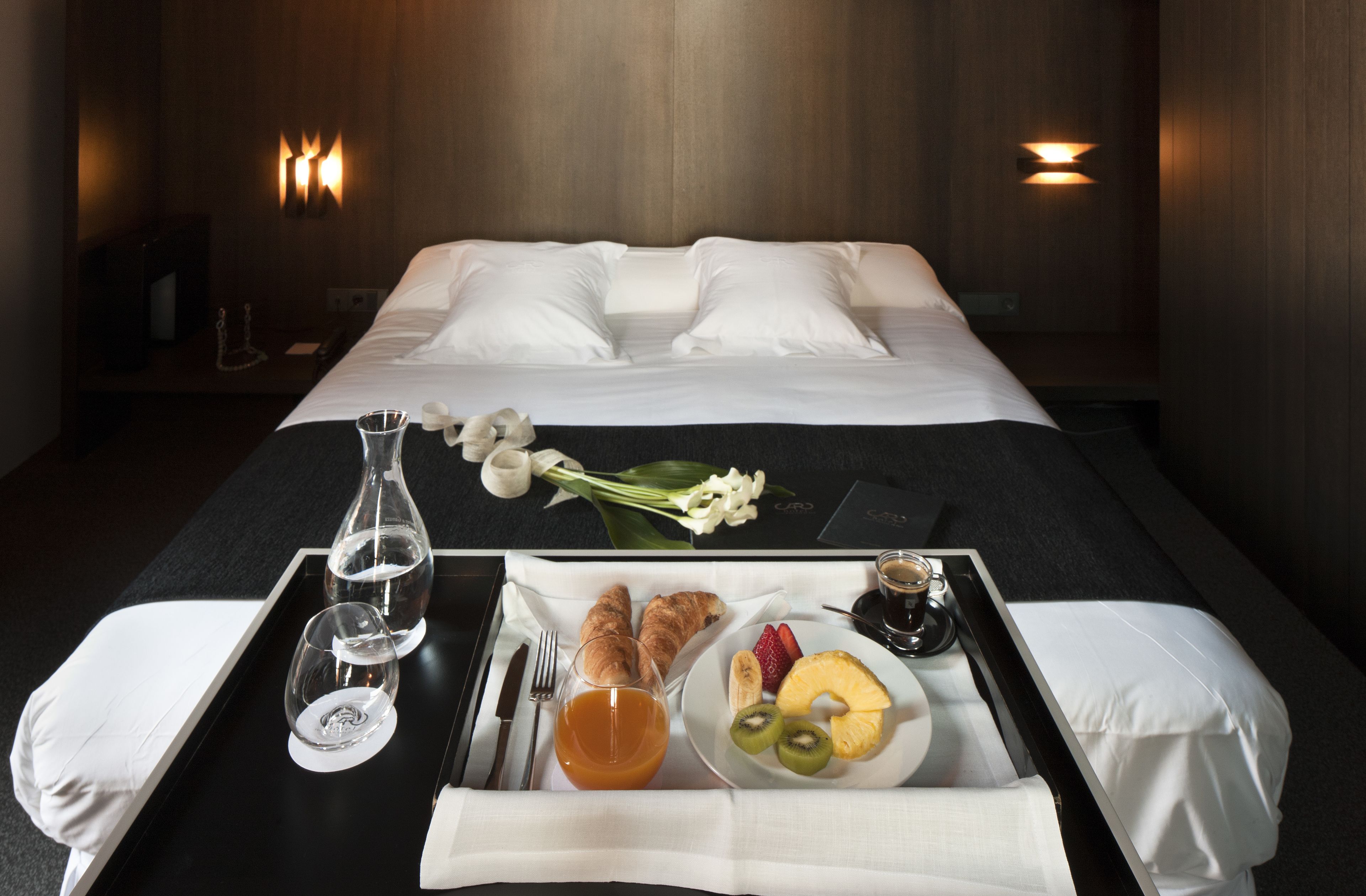 Una cama bien hecha con sábanas blancas, una bandeja con desayuno que incluye croissants, fruta y jugo, y un florero con flores blancas.