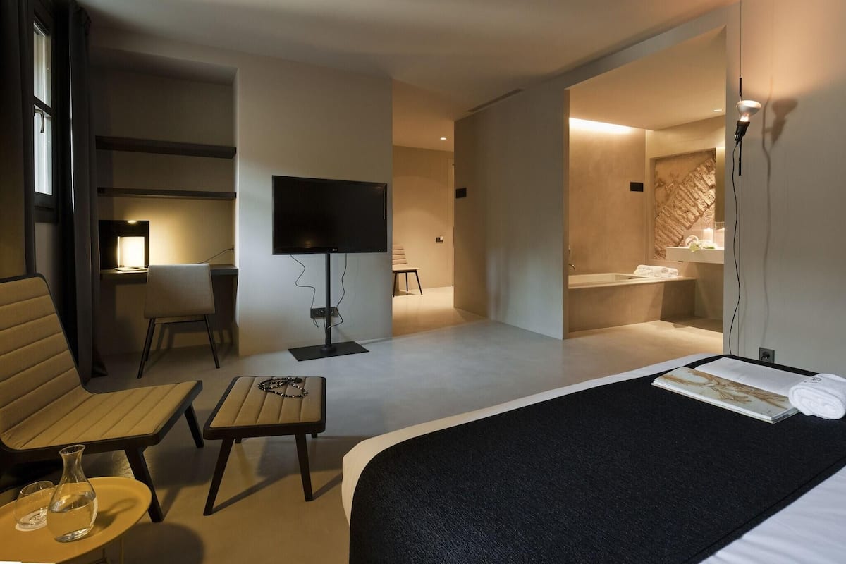 A hotel room with a bed, a TV, a desk with a chair, a small table with a vase and glasses, and a bathroom with a sink and mirror.