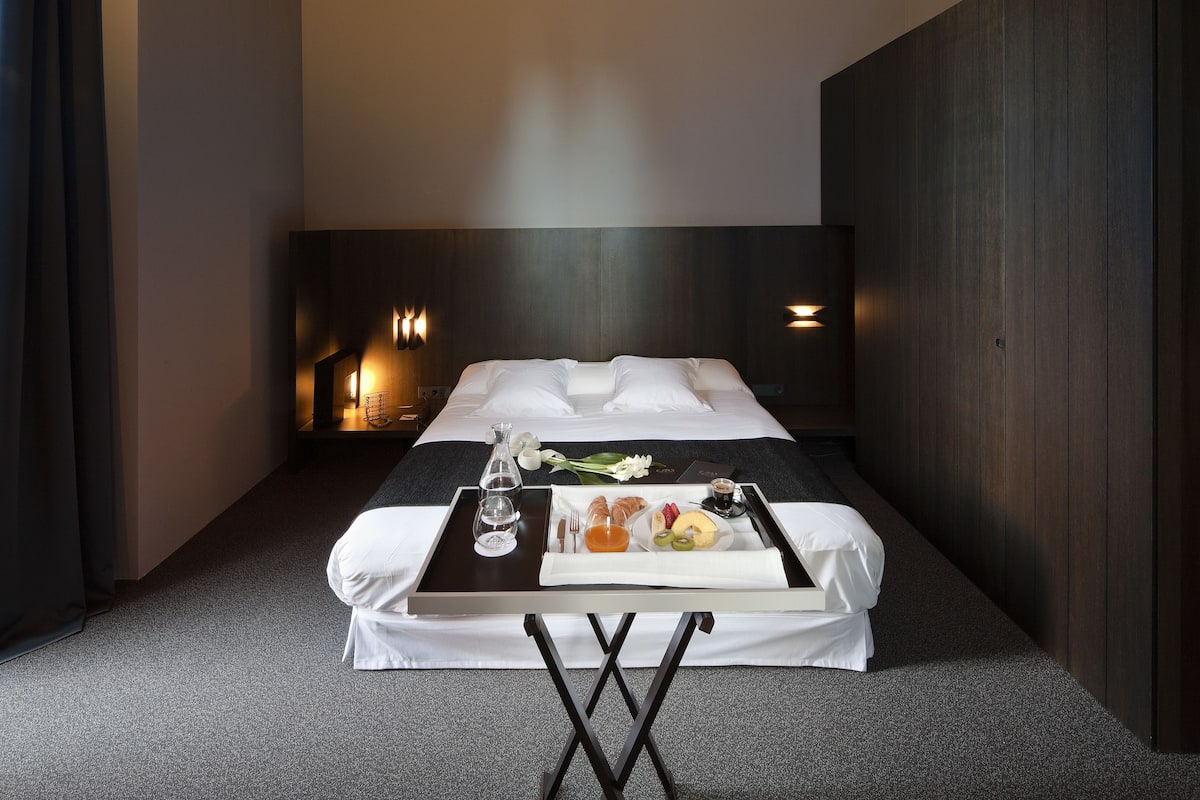 Una habitación de hotel con cama, una mesita con alimentos para el desayuno, una mesita de noche con lámpara y un techo decorado.