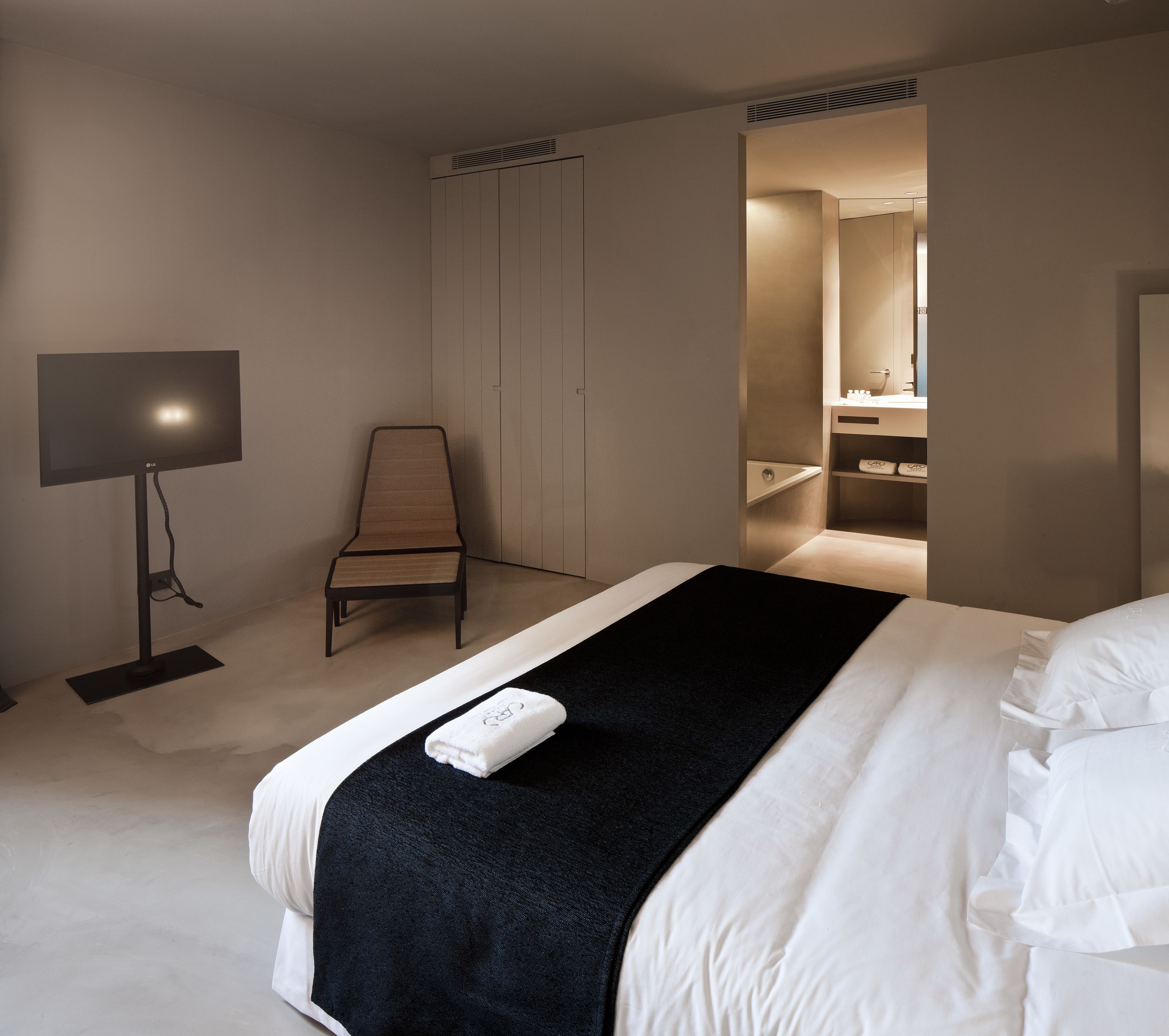 A modern hotel room with a bed, a chair, a TV, and a bathroom visible through an open door.