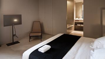 A modern hotel room with a bed, a chair, a TV, and a bathroom visible through an open door.
