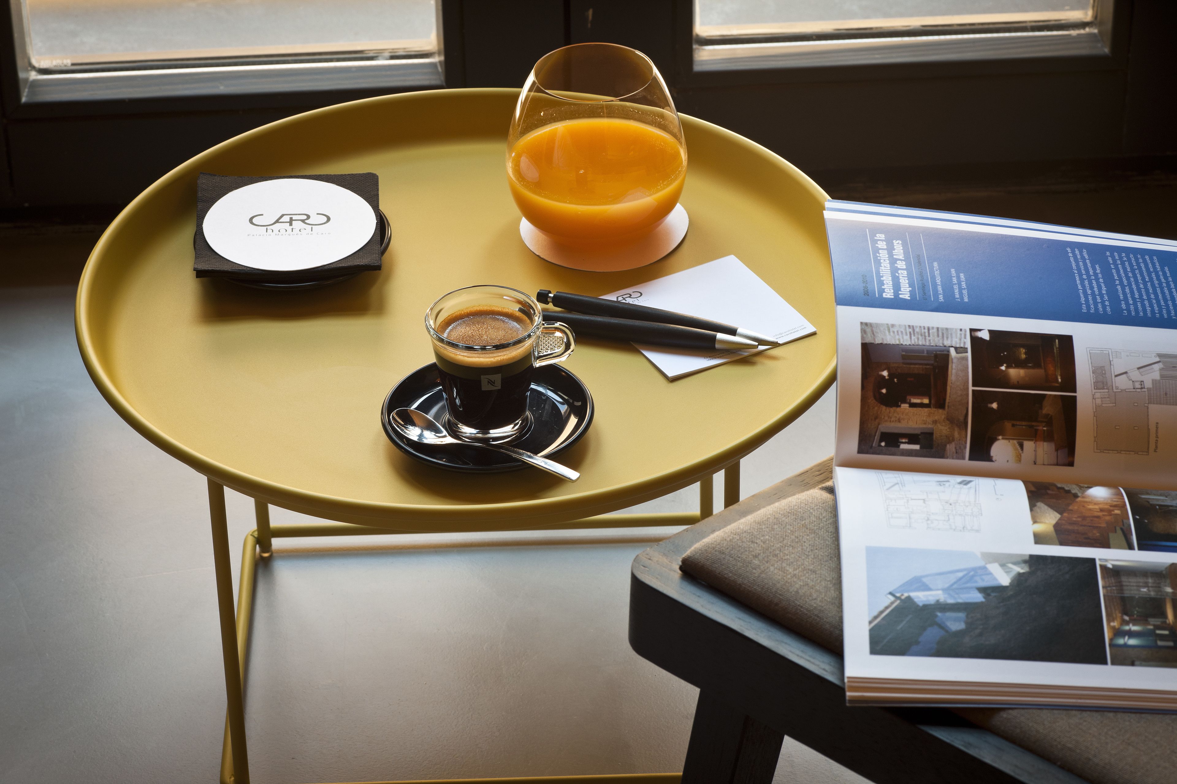 A yellow table with a coffee cup, a glass of orange juice, a pen, and a booklet.