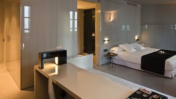Une chambre d’hôtel moderne avec un grand lit, un bureau et une chaise. On y trouve une bibliothèque remplie de livres et une lampe sur le bureau.