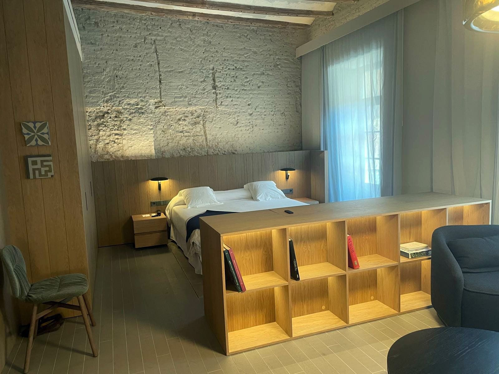 Quarto de hotel com cama, mesas de cabeceira, um aparador de madeira, uma cadeira e uma janela grande com cortinas.